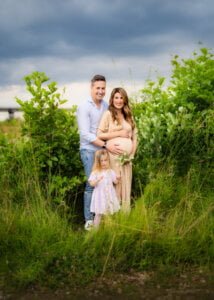Expecting parents with their daughter standing in a green meadow during a maternity photo shoot in Nottingham