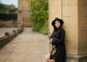 Professional portrait of Nottingham family photographer Marta Szulc standing at Newstead Abbey with camera, wearing a black hat and floral dress during an outdoor branding photoshoot in Nottinghamshire.