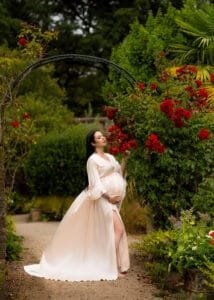 Elegant outdoor maternity photoshoot in Nottingham featuring a pregnant woman in a flowing cream dress surrounded by red roses in a garden